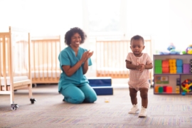 A teacher claps for a toddler as she walks towards a toy.