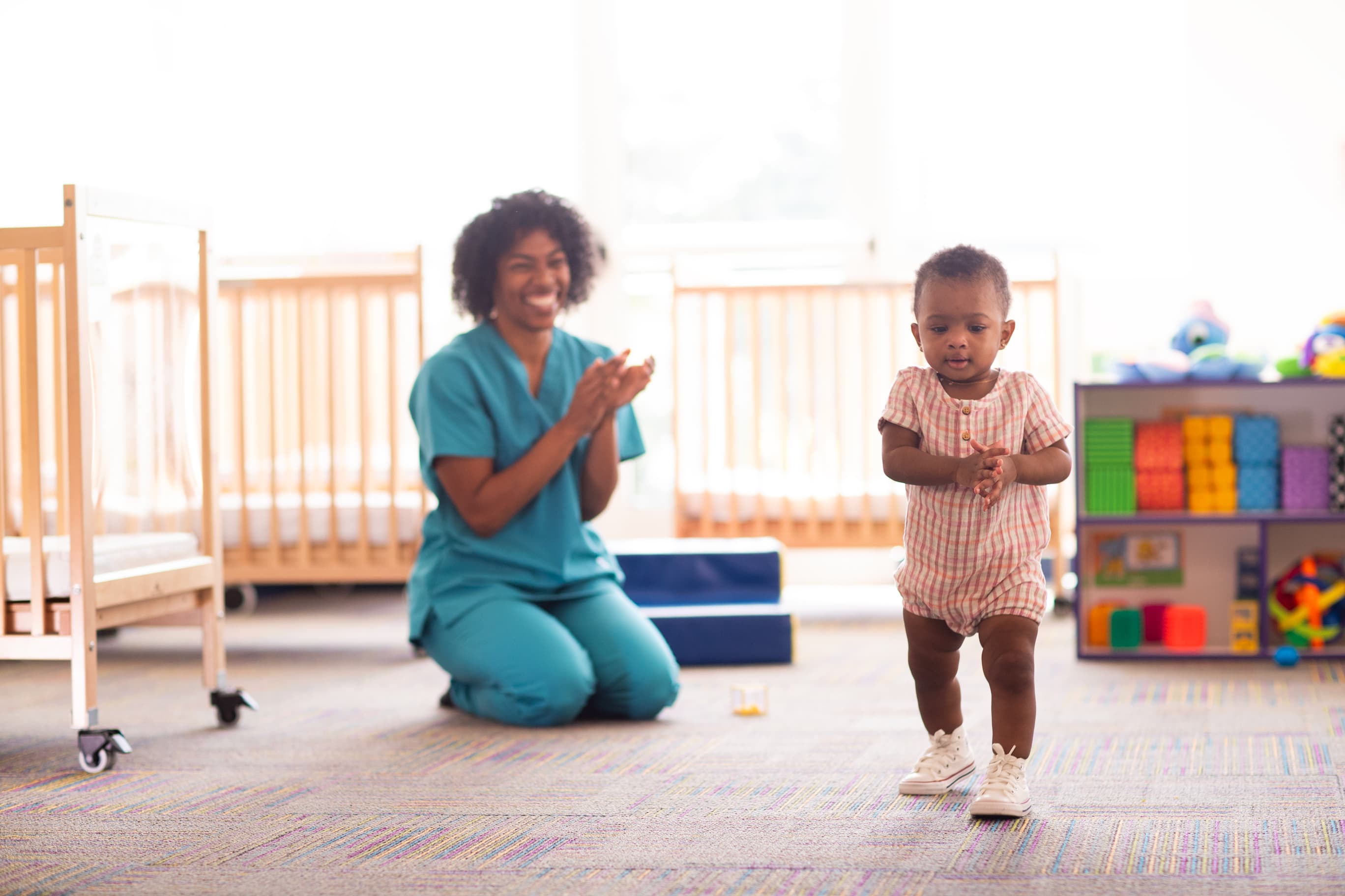 A teacher claps for a toddler as she walks towards a toy.